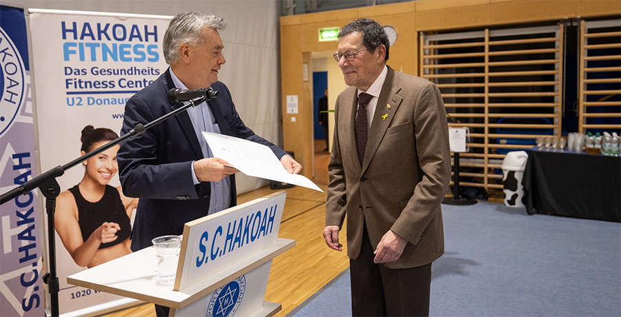 Sportminister Werner Kogler übergibt Urkunde an Professor Paul Haber in einem Turnsaal im S.C. Hakoah